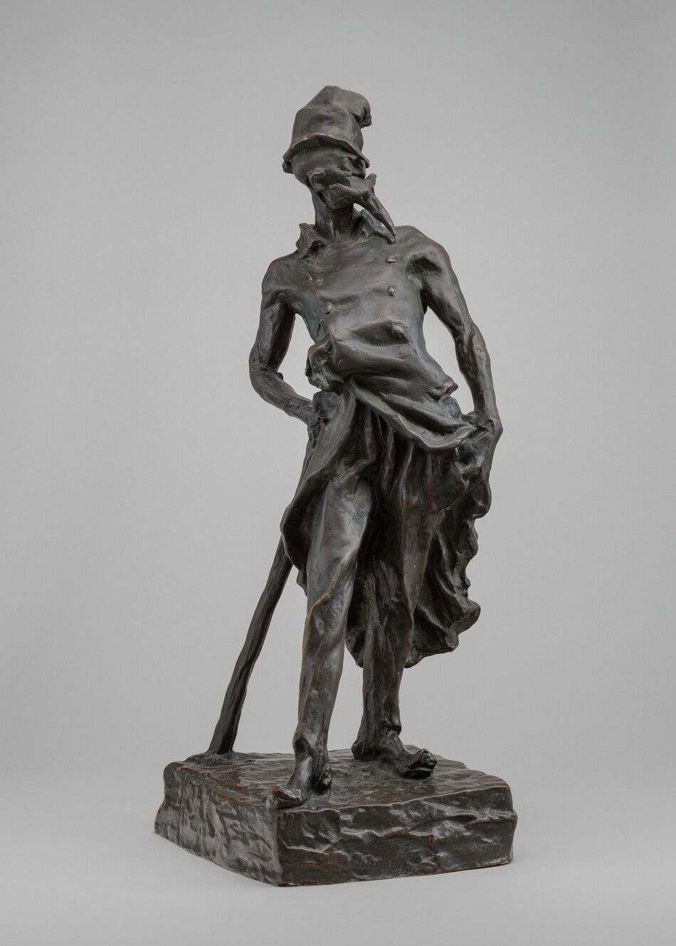 This free-standing bronze sculpture shows a thin man wearing a tall, crumpled hat, an ill-fitting coat, and worn shoes, standing and leaning on a long cudgel resembling a cane. His body faces us in this photograph, and he looks over his shoulder to our right in profile. His goatee is so exaggerated that it creates a beak-like form reaching almost to his chest beneath an upward sweeping mustache that is wider than his face. He has a bull-like nose and bulging eyes, and he might be bald. The top of his tall hat seems to have been partially crushed, and it curves away from us. His double-breasted, knee-length coat has a high collar, and it sweeps open over his hips. His left hand, on our right, holds the coat open so he can slip that hand into his pants pocket. He holds and leans onto the long cudgel on our left so his hips sway to our right. His long pants cover thin legs, and his toes poke through the front of his tattered shoes. The low, square base he stands on is textured as if roughly modeled before being cast. The sculpture is shown against a fog-gray background.