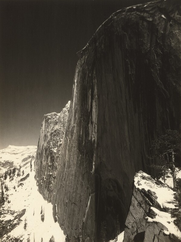 A tall, almost sheer rock formation soaring high above snow-capped rocky peaks nearly fills this vertical black and white photograph. The rock formation is rounded along the top and continues off the top right corner and right edge of the composition. A jagged ridge dips down like a narrow wing to our left, and tall trees on the mountain peaks are made minuscule by the formation, the surface of which is textured with long vertical streaks. The sky in the upper left corner of the photograph is steely gray behind a thin ridge of white snow along the curved top of the formation.
