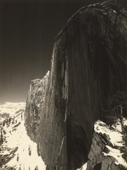 A tall, almost sheer rock formation soaring high above snow-capped rocky peaks nearly fills this vertical black and white photograph. The rock formation is rounded along the top and continues off the top right corner and right edge of the composition. A jagged ridge dips down like a narrow wing to our left, and tall trees on the mountain peaks are made minuscule by the formation, the surface of which is textured with long vertical streaks. The sky in the upper left corner of the photograph is steely gray behind a thin ridge of white snow along the curved top of the formation.