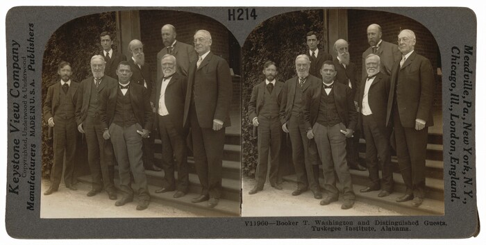 The image is a sepia-toned photograph of a group of men standing together outdoors. They are arranged in a semi-grouped formation on the steps of a building. The man at the center, likely Booker T. Washington, has short hair and is clean-shaven. He wears a three-piece suit and holds a small book or notebook. Other men of varying ages surround him, wearing dark suits with waistcoats and ties. Some individuals are holding hats. The background shows a brick building partially covered by climbing ivy.