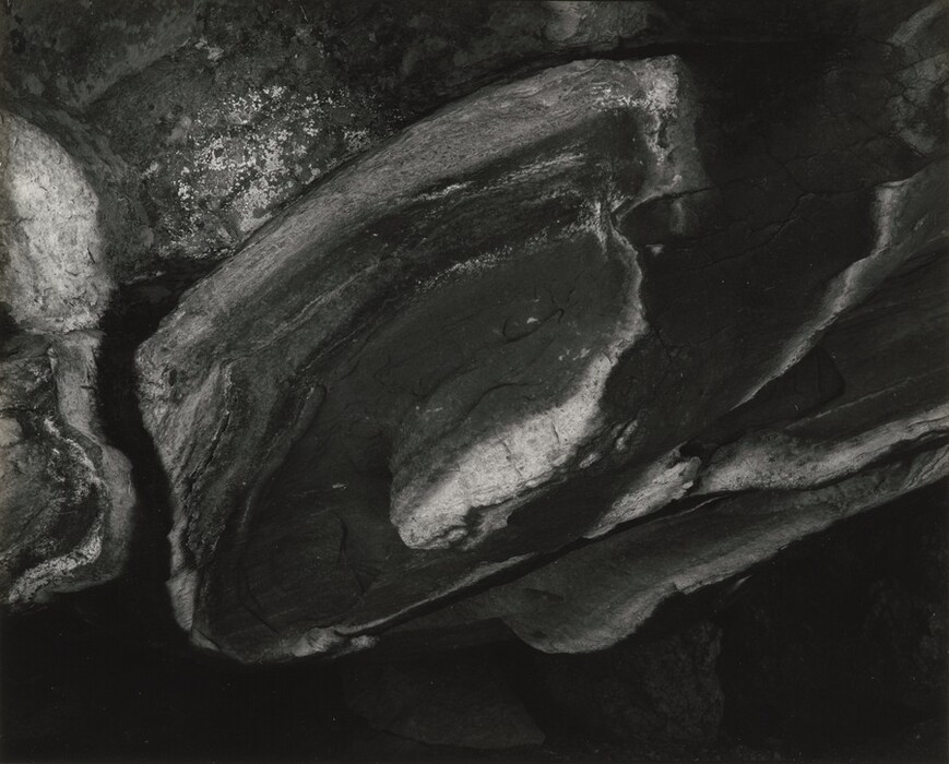 The image shows a pattern of curved, overlapping forms in various shades of light and dark, creating elliptical and oblong shapes. The composition resembles layers of rock or granite with textures suggesting a rugged landscape. The play of shadows and highlights gives the impression of a rough surface, resembling boulders or mountainsides.
