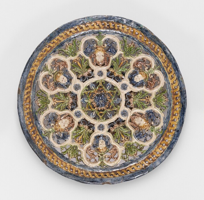 The sculpture is an intricate, circular relief with a detailed pattern resembling a Gothic rose window. The design includes several human faces or busts symmetrically placed around the piece, each nested within a clover-shaped border. Made from glazed ceramic, the sculpture features a rich color palette of deep blues, pastel greens, creamy whites, and golden yellows. The ornate piece has various floral and geometric motifs creating a complex interplay of shapes and patterns. The relief elements stand out against the flat base of the sculpture.