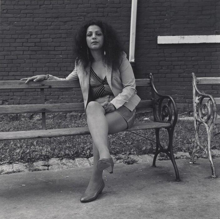 The image depicts a woman sitting on a wooden bench. She has her upper body visible, with one leg crossed over the other. She is leaning back, resting her left arm on the backrest and her right hand on her knee. She has curly hair, a strong facial structure, wearing a blazer over a patterned top, shorts, sheer stockings, and high-heeled shoes. She is wearing large earrings and possibly a bracelet. The background includes a brick wall with white piping, grass, and a concrete path.