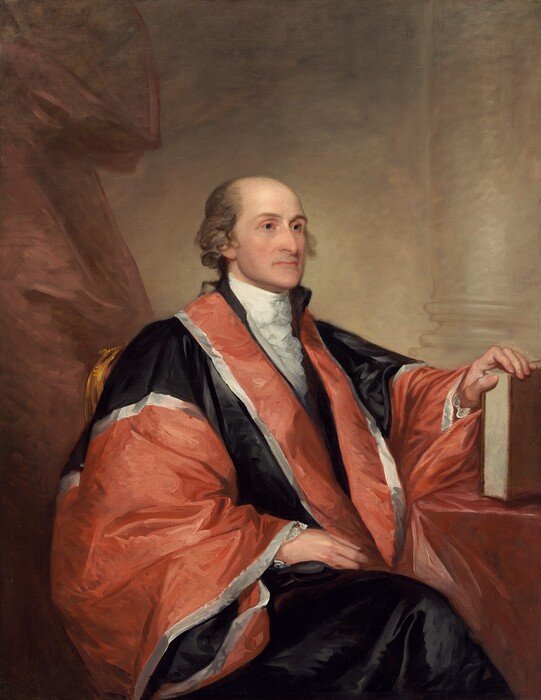 Shown from the knees up, a balding, pale-skinned man wearing a voluminous rust-red and black robe sits angled to our right in this vertical portrait painting. The man is clean-shaven, and tufts of ash-brown hair curls around his ears. He looks to our right with gray eyes under slender brows. The tip of his long nose comes to a prominent point, and his wide, thin, pale pink lips are closed. A white, ruffled ascot is tucked into the lapels of a red and black robe edged with silvery white. The fabric has a sheen, suggesting satin or silk. The man’s legs are crossed and covered with inky-black fabric. The man’s right hand, to our left, rests in his lap, and the other rests on the edge of a book propped upright on a table covered in a dusky, dark pink cloth. A muted copper-red curtain hangs to the left, and a column and its base is outlined against the parchment-brown wall behind him. The portrait is loosely painted with some visible strokes, especially in the costume and background.