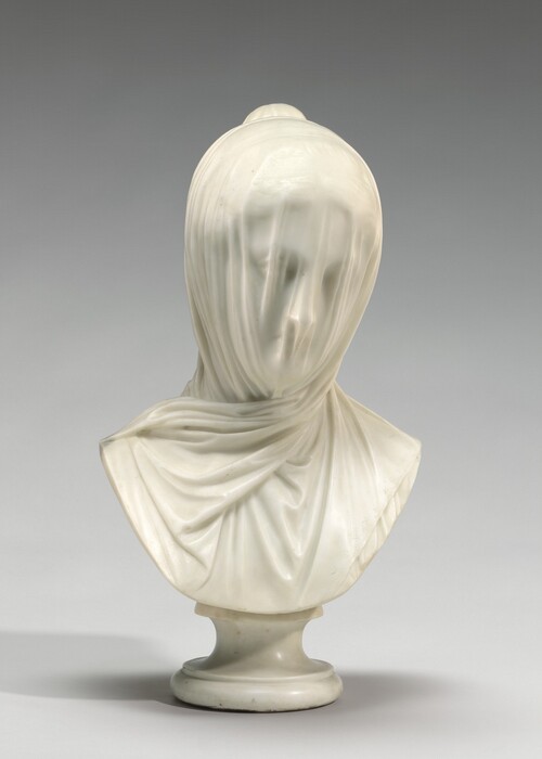 A free-standing, white marble sculpture shows the head and upper chest of a woman whose face is covered by a light veil. In this photograph, the chest is squared toward us, and the woman looks down and to our right. The veil has vertical folds over her face but we can still make out the hollows of her eyes, her straight nose, and her small mouth. Her wavy hair is pulled loosely back into a bun at the back of her crown. The veil gathers on either side of her face and crosses over neck to drape over her chest. The sculpture curves down in a wide U from near her shoulders, and is supported on a white marble pedestal. The background behind the sculpture is pale gray in this photograph.