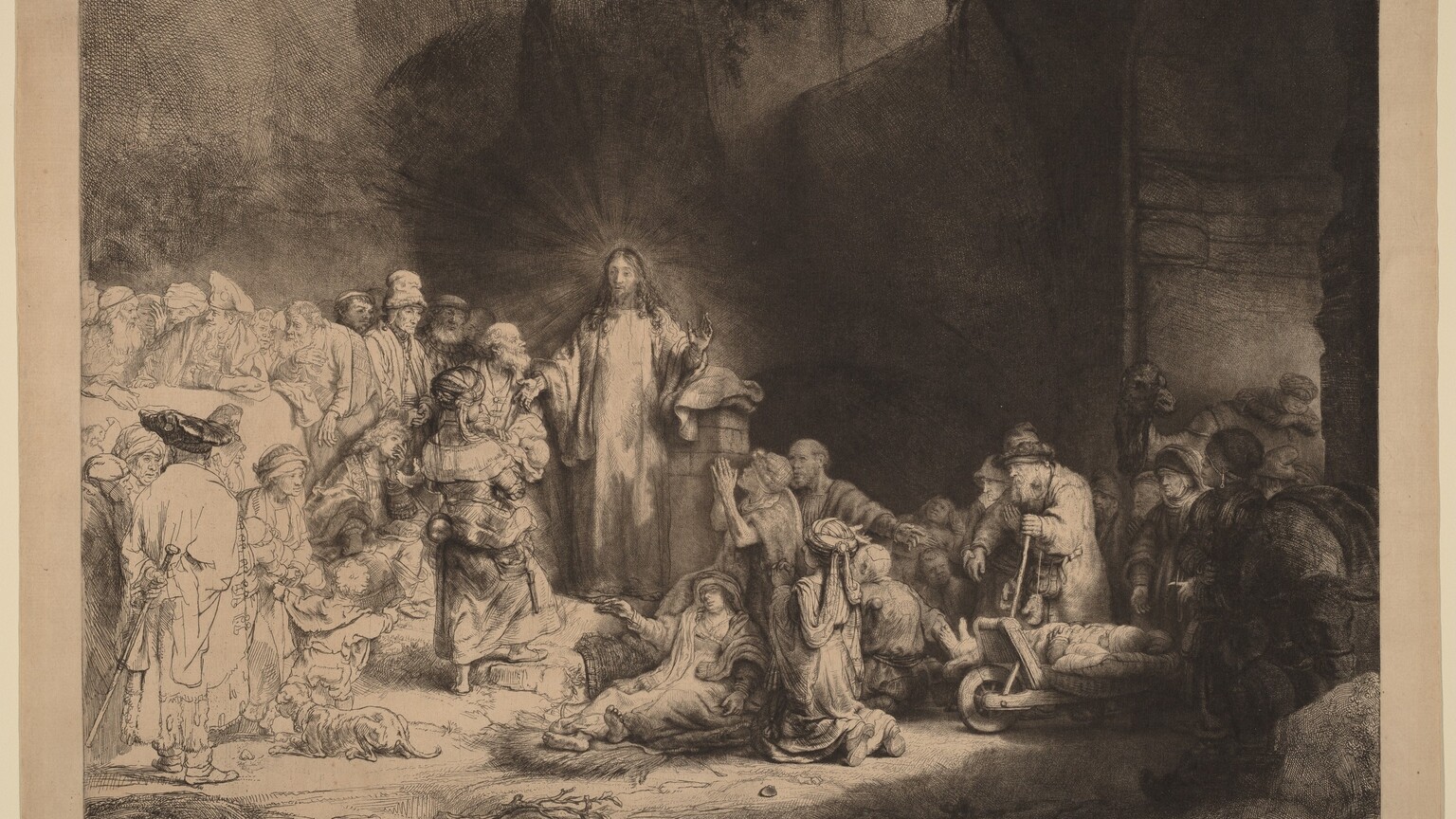 Printed with black lines and deep, velvety shading on tan-colored paper, a man, Jesus, stands at the center of a crowd of more than two dozen people, mostly adults and a few children, in this horizontal etching and drypoint. Jesus is left of center. He stands facing us, but he turns his head to look to our left. Rays of light emanate from behind his head against the towering cave opening that surrounds him. His curly hair falls long over his shoulders, and he holds both hands out. His eyes are lightly incised, giving them an almost blurred look. A crowd of people lie or kneel to our right. Some use crutches or walking sticks, and some are on carts. All the people appear to have pale skin except for one with darker-toned skin to the right in this gathering. That person has a rounded nose, full lips, and wears an earring that gleams brightly in the deep shadows. A camel stands at the far side of this group under an arched opening. The people standing and one person sitting close to Jesus to the left are fully shaded, and those farther left are lightly outlined and sparsely shaded so they appear to be much lighter on the white paper. Close to Jesus are a woman holding a baby, a seated, clean-shaven young man, and several bearded men. Most of those who are along the left edge of the sheet wear turbans or floppy caps.