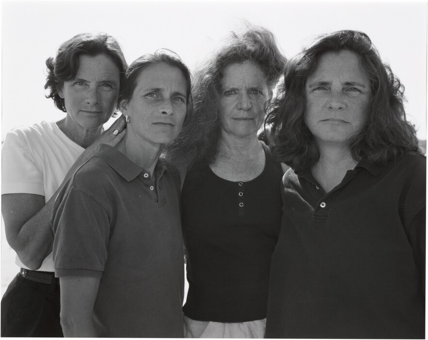 The image shows four women standing closely together from the waist up. They are positioned in a line, with some leaning slightly toward each other. The woman on the far left has short, curly dark hair and is wearing a light short-sleeve top. Next to her, the second woman has long dark hair tied back, with visible facial features including high cheekbones and a determined expression; she is dressed in a dark polo shirt. The third woman from the left has long, curly hair and is wearing a dark top with buttons. The woman on the far right has shoulder-length, wavy hair and is dressed in a dark polo shirt. One woman is seen wearing hoop earrings. The background is a plain, bright sky.