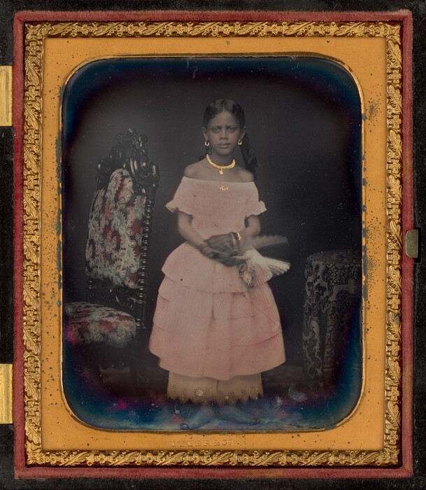 The image depicts a young girl standing upright. She has dark, straight hair with a middle part, falling on her shoulders. She wears a light pink off-the-shoulder dress with multiple tiers and visible underskirt. She accessorizes with a gold-colored necklace, earrings, bracelets, and holds a decorative fan in her hands. Next to her is an ornate chair and a table with a patterned cloth. The background is plain and dark, and the image is framed with intricate gilded details.