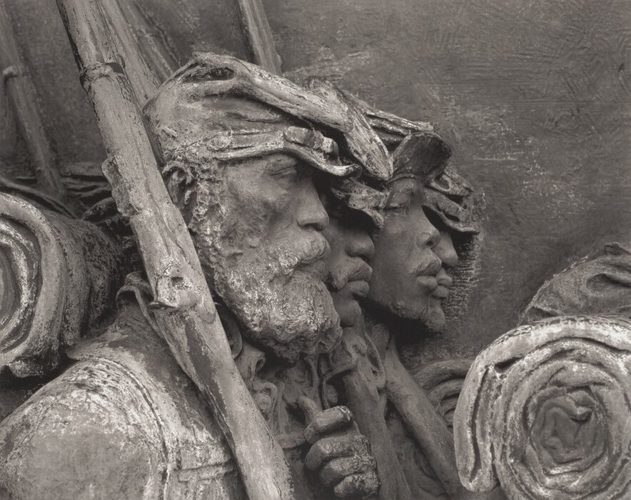 The sculpture depicts three individuals marching in a line. The figures are detailed with facial features and wearing caps, reminiscent of military attire. They carry rolled blankets or bedrolls on their backs and hold onto tools or rifles across their shoulders. The material of the sculpture looks like metal, possibly bronze, giving it a monochrome grayish-brown color. The texture of the sculpture is rough, accentuating the expressions and details of the figures, which are in profile and appear to be engaged in concentrated activity. The figures are closely set, creating a sense of unity and solidarity.