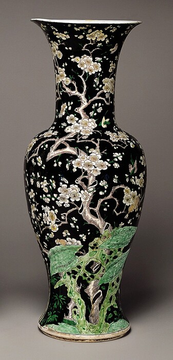 This is a vase featuring a depiction of blooming flowers on a gnarled tree branch. The vase's surface is predominantly black, with detailed white and pale-yellow blossoms and light brown branches. At the bottom are dark green shapes, similar to moss crawling up the side of the tree. The vase appears glossy, and the lip at the top of its narrow neck is flared.