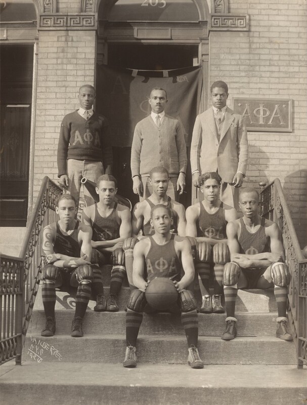 Five young Black men wearing uniforms sit on the front steps of a brick building around a sixth young man, who holds a basketball in this vertical black and white photograph. Three more men stand in a row behind them. They pose on steps leading up to a door covered with a flag and a sign with the Greek letters for alpha phi alpha. The seated men, the players, wear knee pads, knee-high socks, and sleeveless tank tops with the Greek letters. One of the men in the background wears an Alpha Phi Alpha sweater over a shirt and tie. The next wears a sweater jacket and the third a suit jacket, all over shirts and ties. The artist signed this photograph by inscribing the negative, “VANDERZEE N.Y.C. 1926.”