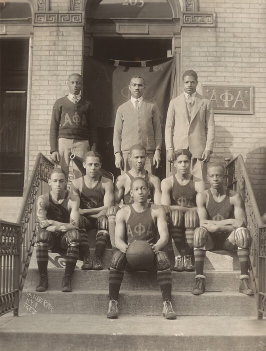 Five young Black men wearing uniforms sit on the front steps of a brick building around a sixth young man, who holds a basketball in this vertical black and white photograph. Three more men stand in a row behind them. They pose on steps leading up to a door covered with a flag and a sign with the Greek letters for alpha phi alpha. The seated men, the players, wear knee pads, knee-high socks, and sleeveless tank tops with the Greek letters. One of the men in the background wears an Alpha Phi Alpha sweater over a shirt and tie. The next wears a sweater jacket and the third a suit jacket, all over shirts and ties. The artist signed this photograph by inscribing the negative, “VANDERZEE N.Y.C. 1926.”