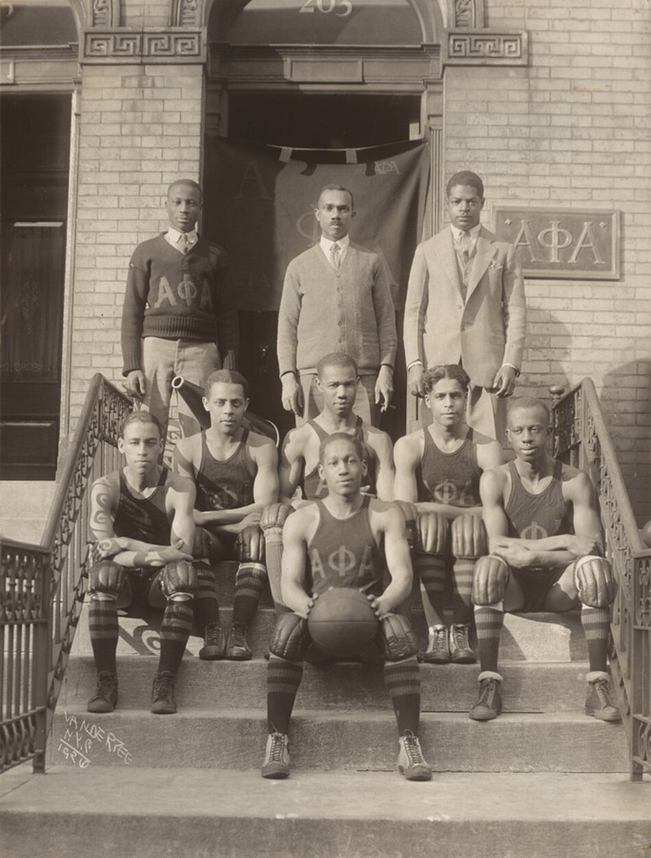 Five young Black men wearing uniforms sit on the front steps of a brick building around a sixth young man, who holds a basketball in this vertical black and white photograph. Three more men stand in a row behind them. They pose on steps leading up to a door covered with a flag and a sign with the Greek letters for alpha phi alpha. The seated men, the players, wear knee pads, knee-high socks, and sleeveless tank tops with the Greek letters. One of the men in the background wears an Alpha Phi Alpha sweater over a shirt and tie. The next wears a sweater jacket and the third a suit jacket, all over shirts and ties. The artist signed this photograph by inscribing the negative, “VANDERZEE N.Y.C. 1926.”