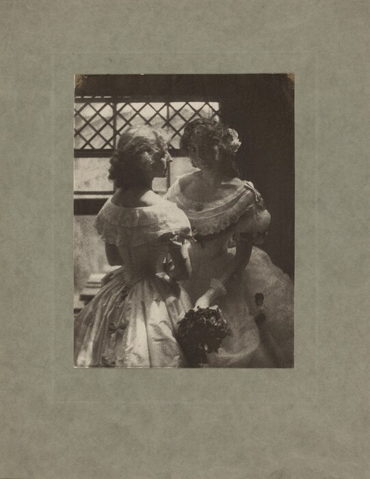 The image shows two women captured in a vertical composition where both are visible from the waist up. They are positioned facing each other in a close manner. The woman on the left is seen in profile with hair styled in soft curls framing her face. The woman on the right is looking slightly down with her head turned towards the other, her hair also curly with a noticeable flower adorning it. Both women wear elegant and elaborate dresses with puffed sleeves and lace detailing. The woman on the right holds a small bouquet of dark flowers in her right hand. They appear to be in an indoor setting, in front of a window with a lattice pattern, casting a soft light into the room. The background is softly out of focus, emphasizing the scene between the two figures.