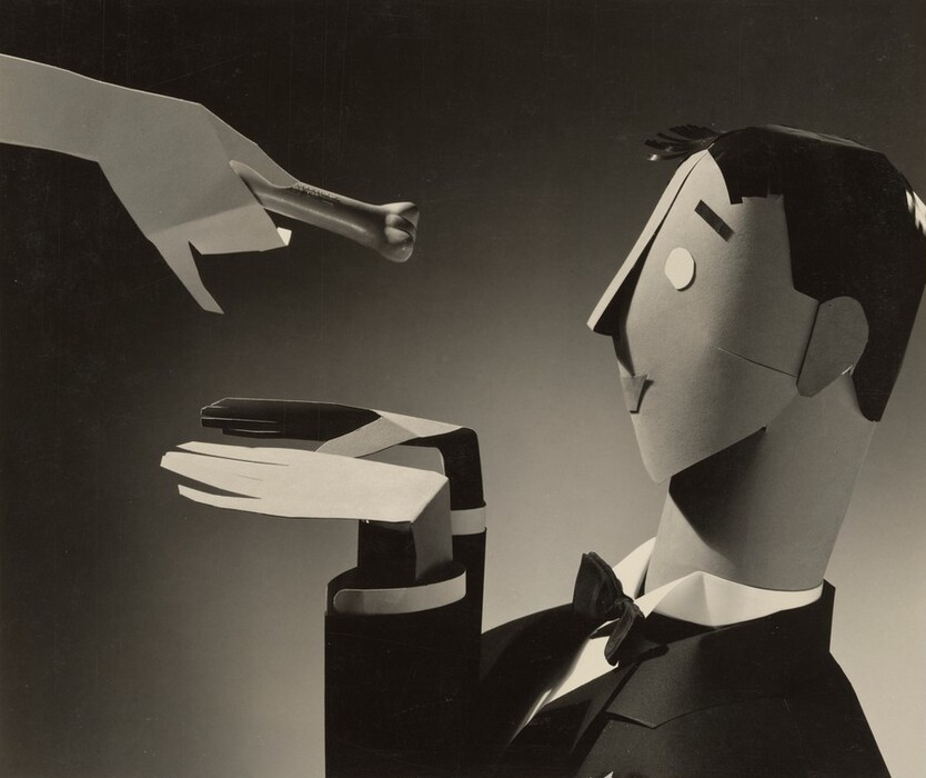 This is a photograph of an abstract composition using paper cut-outs and a lipstick. It shows a paper-crafted hand extending a lipstick towards a cut-out figure of a head and arm, positioned as if ready to receive the object. The head is minimalist in design, with simple shapes and lines suggesting facial features like eyes, nose, and a subtle indication of a mouth. The figure is dressed in a tuxedo, indicated by angular folds resembling a bow tie and contrasting tones implying lapels and collars. The lighting highlights shadows and depth, emphasizing the paper edges and the overall composition's elegance.