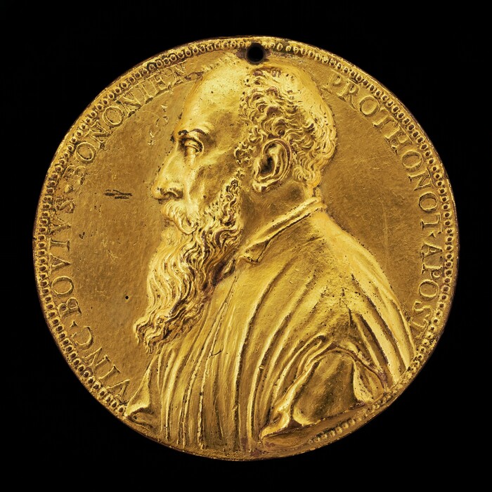The sculpture is a circular medallion featuring the profile of a bearded man facing left. The man has a prominent forehead and a long, flowing beard, with detailed curly hair. He appears to be wearing a robe or tunic with fabric folds. The medallion is a rich golden color, suggesting a gilded material such as brass or gold alloy. Around the edge of the medallion is an inscription in raised letters, and the design indicates attention to detail. The surface of the medallion shows variations in texture, and there is a small hole at the top.