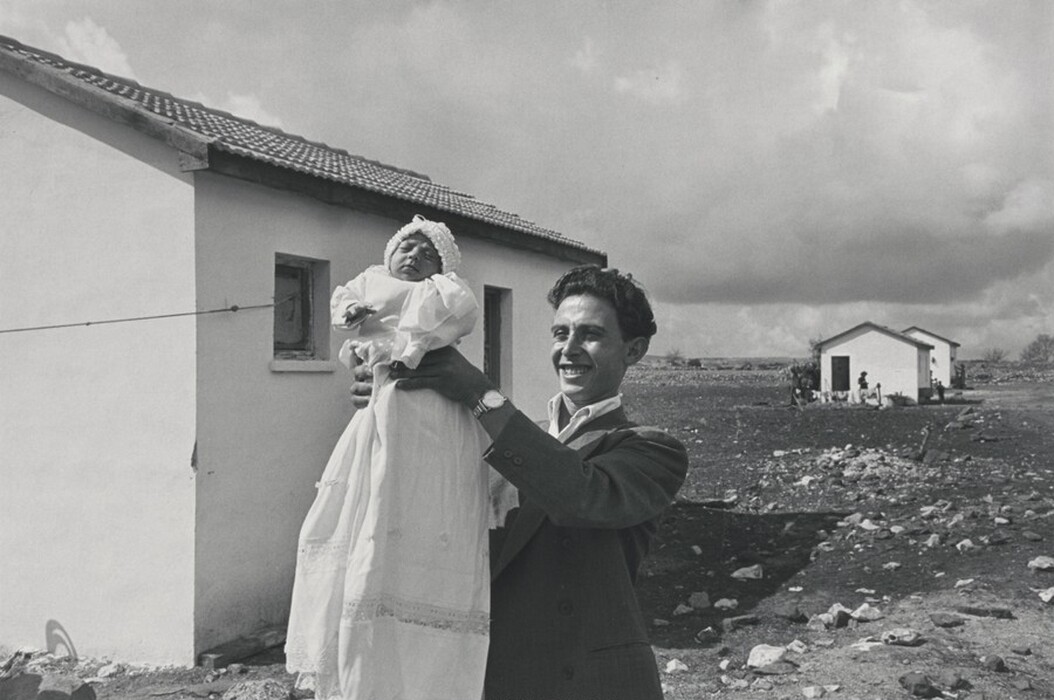 The image features a man holding a baby, both posed in front of two small white houses with tiled roofs. The man is smiling and dressed in a dark suit with a white shirt, and the baby is in a white gown and bonnet, eyes closed and facing upwards. The background includes a clothesline, with a cloudy sky overhead and dirt ground with scattered stones.