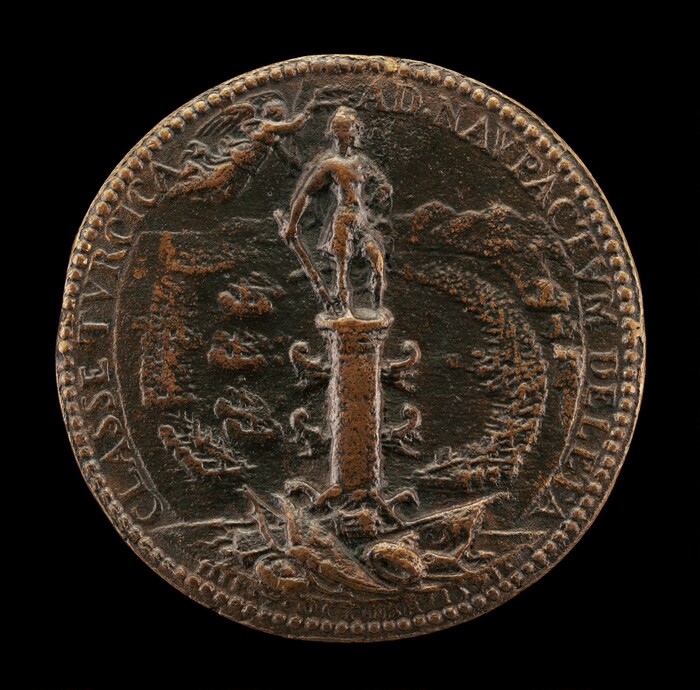 This is a photograph of an ancient coin or medallion. The coin features a central figure standing on a pedestal or column, surrounded by intricate designs and smaller figures. The surface of the coin shows signs of wear and patina. Around the outer edge, there is an inscription whose letters are visible but worn. The central figure appears to be holding an object, and additional small depictions can be seen around the pedestal. The artistry of the coin reflects a high level of craftsmanship typical of historical minting, with attention to detail in both the figure and the surrounding designs.