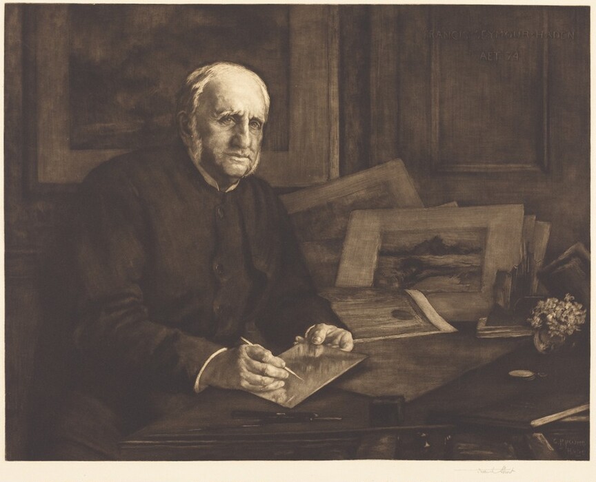 The image shows an older man sitting at a desk. He is turned slightly to his right, with his right arm resting on the surface and his left hand holding a pencil over a sheet of paper or board. He has a prominent nose, deep-set eyes, and a thoughtful expression, as well as short, mostly white hair that is receding at the temples. He is wearing a dark coat or tunic. On the desk are sketches, prints, and paper materials, along with a pot of flowers and a rectangular object, possibly a magnifying glass. The background features wooden paneling and a framed picture, creating an office or study setting.