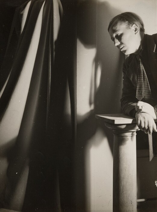 This grayscale photograph shows the upper body of a person leaning on a short column, with their head tilted and their face in profile looking to the left. They have pale skin and their hair is short and combed. They are wearing a dark suit with a dotted tie, and their hands rest on the column. The background includes a curtain and dramatic shadows on the wall to the left.