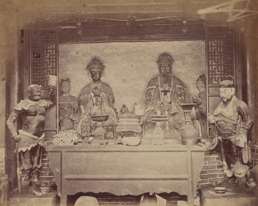 The image depicts a group of sculptures arranged within an ornate setting. The wooden table in the foreground is adorned with various objects. The subjects of the sculptures are various figures. The central figures are two larger-than-life statues seated behind the table. They are flanked on either side by smaller, more intricately detailed sculptures. The sculpture on the left holds a paper or document, while the one on the right is poised with a staff or tool. The figures are crafted from a material that appears aged, possibly wood or stone, with a dark, earthy color patina. The details suggest clothing with ornate patterns and textures. The setting includes lattice screens on either side. Overall, the sculptures collectively present a tableau reflecting cultural or traditional motifs evident in their attire and posture.