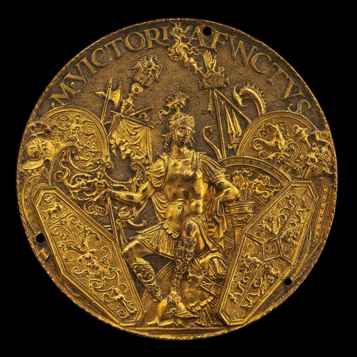 The sculpture is circular and resembles a medallion or relief. It features a seated figure of a warrior or deity in a commanding pose. The subject is adorned in classical armor with intricate detailing showing a mix of muscular and fabric textures. The warrior holds a shield with elaborate designs and is surrounded by ornate elements, including a helmet and spears. The entire piece is crafted from a material with a gold appearance, giving it a metallic sheen. The sculpture's circular form and small holes suggest it may have been a decorative attachment.