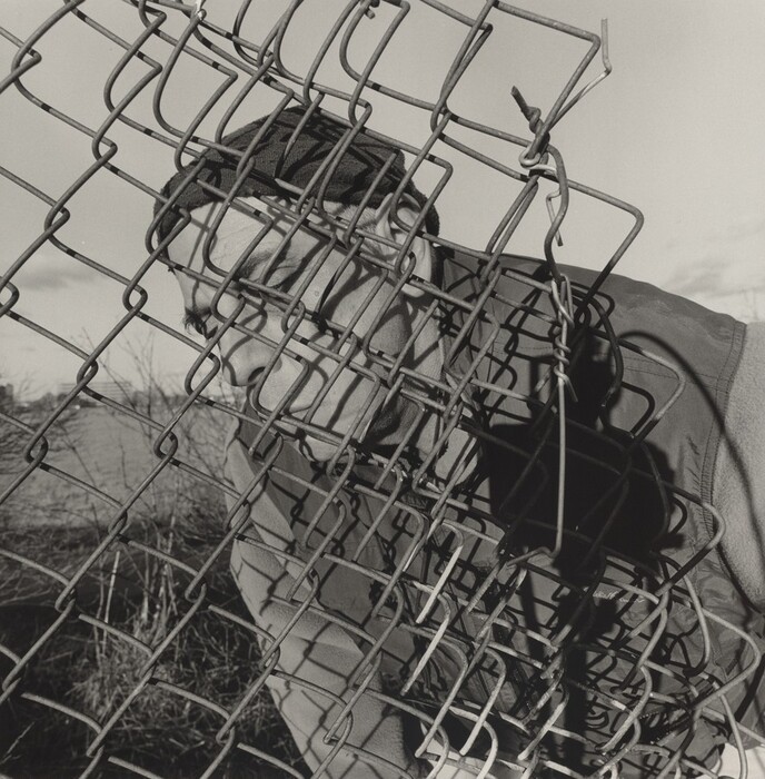 The image depicts the upper part of a person's body, including their head and shoulders. The individual is seen bending forward with a metal mesh fence partially obscuring their face. The person has short dark hair, potentially wearing a beanie, and their facial expression appears concentrated with their eyes partly closed or squinting. They are dressed in a thick, textured jacket or coat. The background shows an outdoor setting with vegetation visible through the fence. The sky is clear with a few clouds. Shadows from the fence create a layered visual effect on the person's face and clothing.