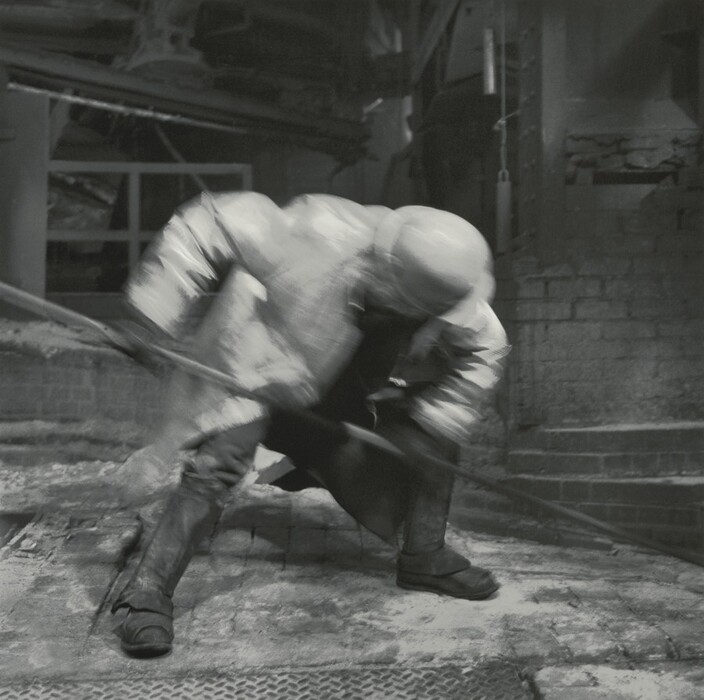 The photograph depicts an industrial scene with a blurred figure in a factory or workshop setting. The image captures motion and a sense of urgency, with machinery and brick walls visible. The blur effect creates a dynamic energy in the scene.