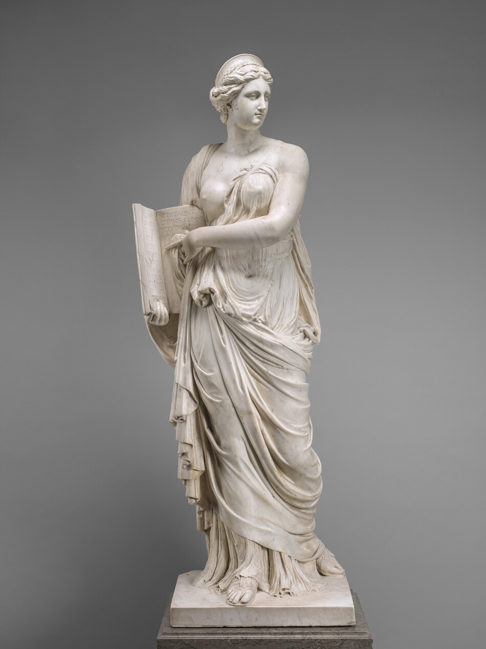 A woman carved from cream-white marble stands holding an open book to which she points in this free-standing sculpture. In this photograph, her body faces us but her head turns to our right. She has a long nose, small mouth, and rounded chin. Her wavy, upswept hair is topped by a semi-circular headdress. Her voluminous gown clings to her torso, leaving one breast exposed. The fabric falls in deep folds around her legs and sandalled feet. Her left shoulder, on our right, is bare. A band crosses her chest from that shoulder to her waist. She holds the tall book in her right hand, on our left, braced against that hip. Her other hand points to the words inscribed in Latin on its pages. It reads, “CALLIOPE REGI NA,HOMINVM, DIVVMQVE VO LVPTAS CARMINIS HE ROI NVMERIS FVLGENTIA SIGNIS AGMINA, BEL LANTVMQVE ANIMOS, ET P RAELIA CAN TO, INCLYTAQVE AETERNAE COMMI TO. NOMINA FAMAE.” She stands on a square base supported on a fawn-brown, veined marble plinth. The background behind her is nickel gray.