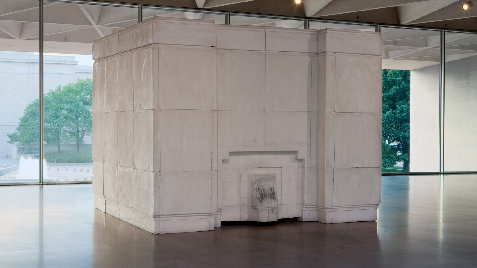 Set in a gallery against a row of windows, a free-standing, white plaster cube, nearly reaching the ceiling, angles back and away from us slightly to our right. The cube is made of blocks that fit together so the seams are visible. The side to our left is smooth. A short, tongue-like projection at the bottom center of the face to our right is surrounded by an inverted, squared off U arching over it. Though not obvious in this photograph, the artist created this work by filling an empty room with plaster. The tongue-like shape is the inside of a fireplace, and is lightly blackened where it lined the interior. The inset paneling in the cube would have been the room's mantlepiece, and inset bands along the floor and top of the cube would have been the baseboards and crown molding. The cube sits on a polished stone floor, and the ceiling of the room in which it sits is made of hollow triangular coffers. Opposite us, trees and a building are seen through the wall of windows behind the cube.