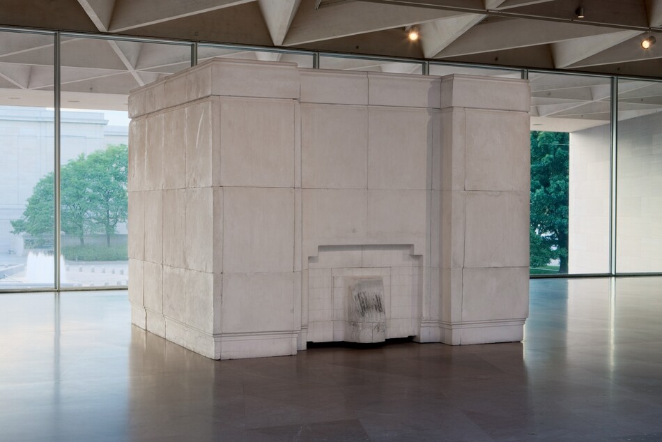 Set in a gallery against a row of windows, a free-standing, white plaster cube, nearly reaching the ceiling, angles back and away from us slightly to our right. The cube is made of blocks that fit together so the seams are visible. The side to our left is smooth. A short, tongue-like projection at the bottom center of the face to our right is surrounded by an inverted, squared off U arching over it. Though not obvious in this photograph, the artist created this work by filling an empty room with plaster. The tongue-like shape is the inside of a fireplace, and is lightly blackened where it lined the interior. The inset paneling in the cube would have been the room's mantlepiece, and inset bands along the floor and top of the cube would have been the baseboards and crown molding. The cube sits on a polished stone floor, and the ceiling of the room in which it sits is made of hollow triangular coffers. Opposite us, trees and a building are seen through the wall of windows behind the cube.