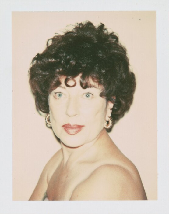 The image shows a head-and-shoulders portrait of a woman. She is positioned slightly turned to her left with her head turned towards the camera. Her facial features include light-colored eyes and red lipstick. Her hair is styled in voluminous, curly waves and is dark in color with a notable curl on her forehead. She is wearing a strapless garment with visible bare shoulders and has hoop earrings as jewelry. The background is a plain, soft peach color.