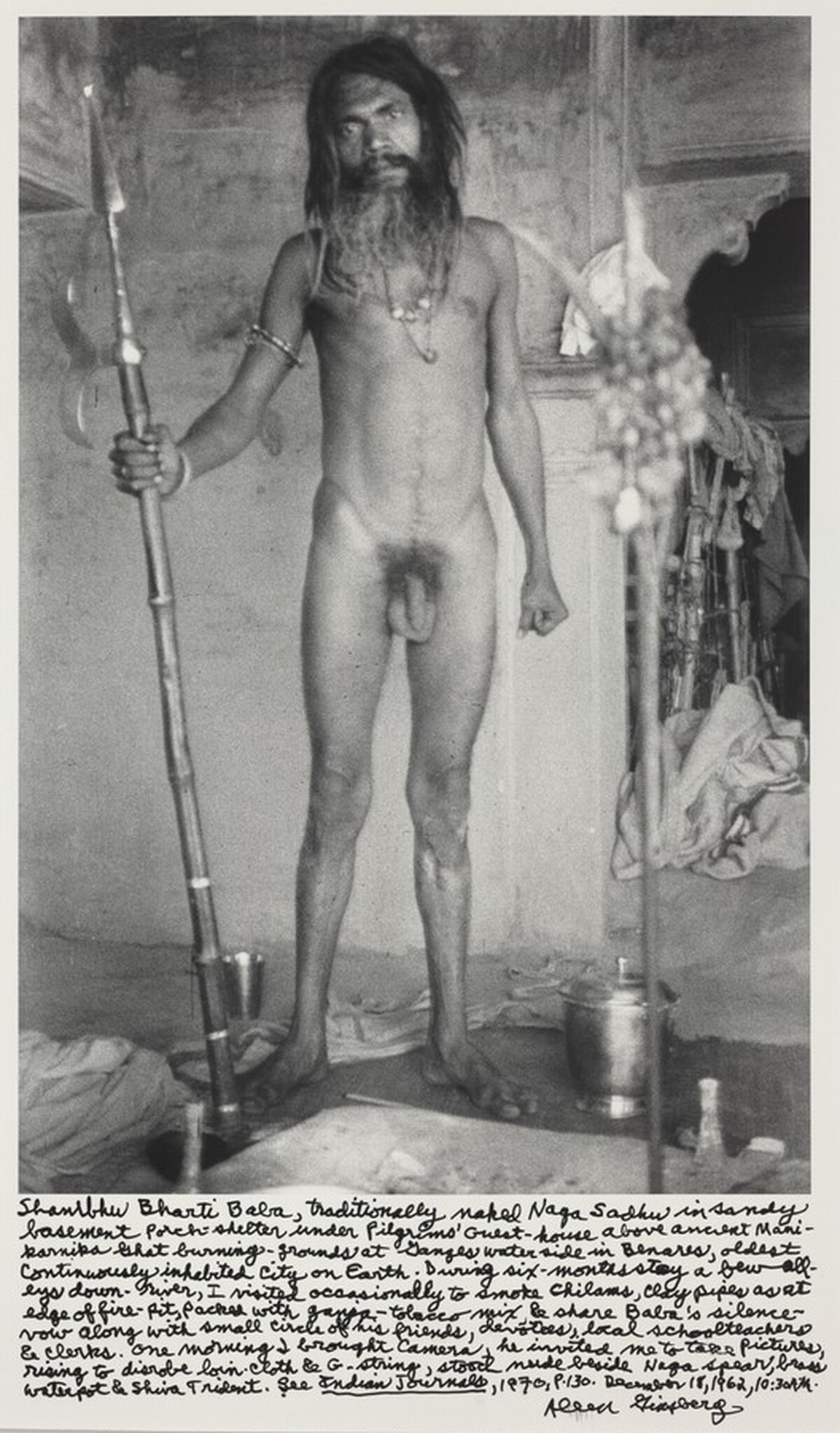 Shambhu Bharti Baba, traditionally naked Naga Sadhu in sandy basement porch-shelter under Pilgrims' Guest-house above ancient Manikarnika Ghat burning-grounds at Ganges waterside in Benares, oldest cintinuously inhabited city on Earth. During six-months stay a few alleys down-river, I visited occasionally to smoke chilams, clay pipes as at edge of fire-pit, packed with ganja-tobacco mix & share Baba's silence vow along with small circle of his friends, devotees, local schoolteachers & clerks. One morning I brought camera, he invited me to take pictures rising to disrobe loin-cloth & G-string, stood nude beside Naga spear, brass waterpot & Shiva Trident. See _Indian Journals_, 1970, p.130. December 18, 1962, 10:30 AM.