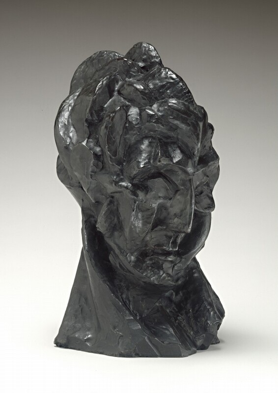 A woman’s face and neck are roughly modeled in this dark green, freestanding bronze sculpture. In this photograph, her face is angled to our right. She looks down, chin tucked back into her neck. She has deep-set eye sockets, a thin, blade-sharp nose, and her small mouth is closed, the corners downturned. Light from our left gleams off some of the surfaces, especially in the choppy hair and along the edge of her nose. A long diagonal ridge on the right side of her neck, our left, suggests a tendon stretching as she turns her head. Her neck acts as the sculpture's base. The background lightens from pale gray along the top to white along the bottom, where the sculpture casts a faint shadow to our right.