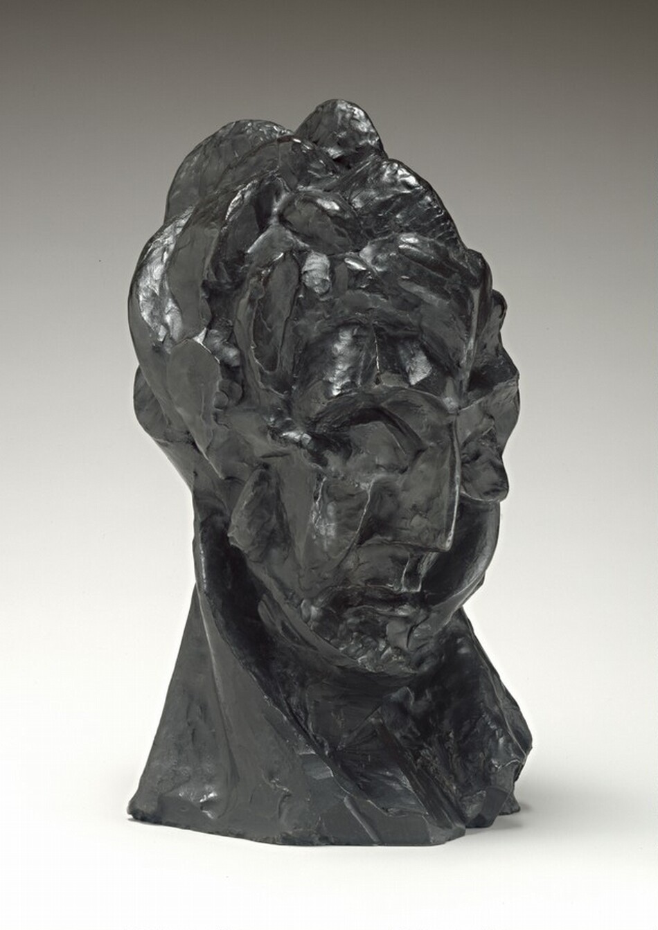 A woman’s face and neck are roughly modeled in this dark green, freestanding bronze sculpture. In this photograph, her face is angled to our right. She looks down, chin tucked back into her neck. She has deep-set eye sockets, a thin, blade-sharp nose, and her small mouth is closed, the corners downturned. Light from our left gleams off some of the surfaces, especially in the choppy hair and along the edge of her nose. A long diagonal ridge on the right side of her neck, our left, suggests a tendon stretching as she turns her head. Her neck acts as the sculpture's base. The background lightens from pale gray along the top to white along the bottom, where the sculpture casts a faint shadow to our right.