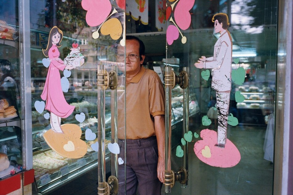 This photograph shows a man standing behind a partially open glass door, with half of his body obscured. He is seen from the knees up, looking directly us. He has short, dark hair, wrinkled light-brown skin, and is wearing glasses. His attire includes a tan polo shirt and dark pants. The glass door is adorned with colorful illustrations, including the cartoon figures of a man and a woman with heart shapes surrounding them. In the background, there are displays of various baked goods inside the store. The landscape outside is partially reflected in the glass.