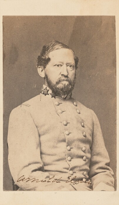 The image depicts a man seated, facing forward with a serious expression. He has wavy hair, a full beard, and a mustache. He wears a military-style coat with stars on the collar. The background is plain.
