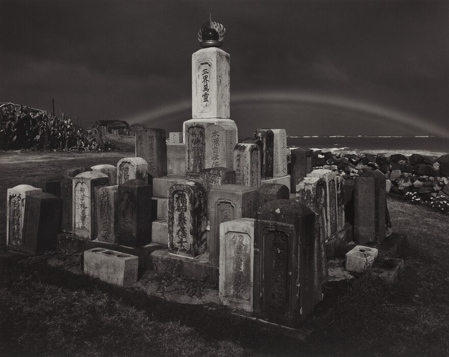 The image shows a low viewpoint looking up slightly, with a horizon near the top third. A curved line in the sky may be a rainbow. The sky is overcast, contrasting with the rough sea. A group of aged grave markers of varying sizes and shapes are in the foreground, with inscriptions visible on some. A stone fence runs parallel to the horizon, with a small structure and foliage in the distance on the left. The color palette is monochromatic, emphasizing textures and contrasts between light and shadow.
