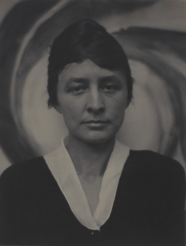Shown from the chest up, a woman faces and looks at us in front of a swirling painting in this vertical black and white portrait photograph. The woman has thick, dark brows over slightly downturned eyes. Her lips are closed, and her dark hair is pulled back. Her dark garment has a white collar creating a narrow V below her throat. She stares steadily out in front of the dark and light-colored spiraling strokes of the painting behind her.