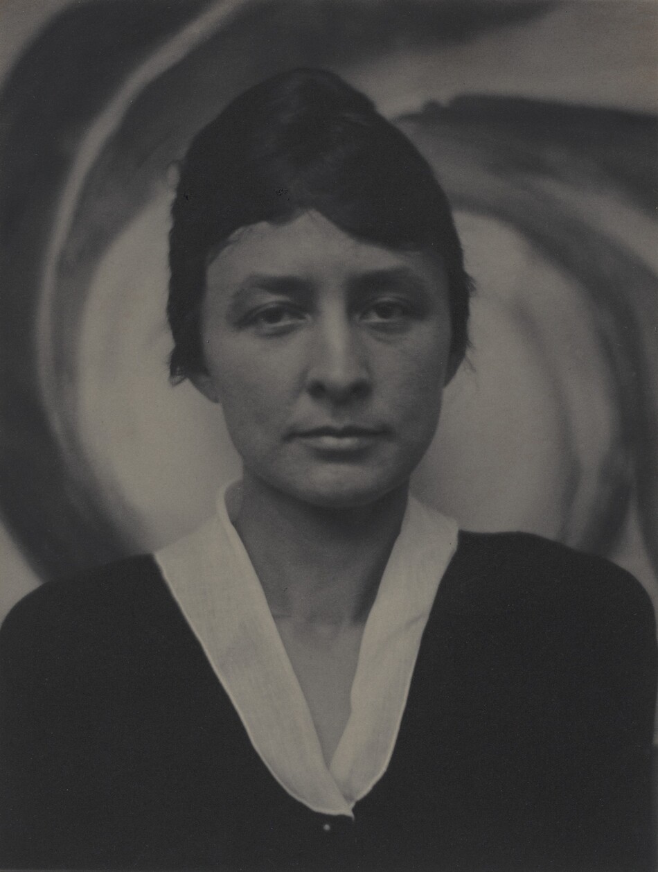 Shown from the chest up, a woman faces and looks at us in front of a swirling painting in this vertical black and white portrait photograph. The woman has thick, dark brows over slightly downturned eyes. Her lips are closed, and her dark hair is pulled back. Her dark garment has a white collar creating a narrow V below her throat. She stares steadily out in front of the dark and light-colored spiraling strokes of the painting behind her.