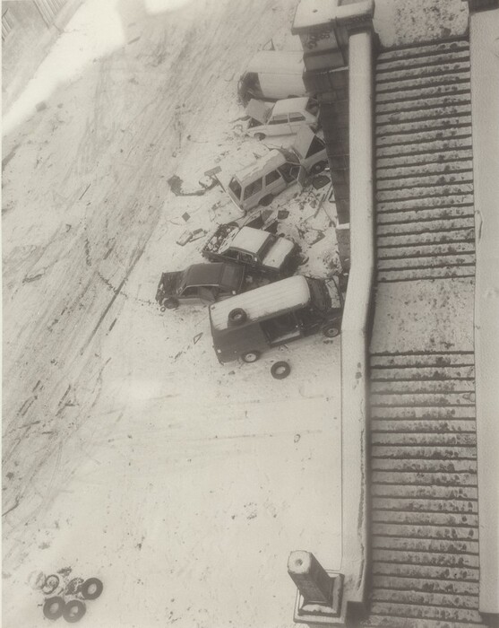 The image shows a snow-covered urban scene from a top-down perspective. Several cars are parked next to a vertical structure that seems to be the edge of a flight of steps. The steps create a repetitive pattern on the right side. The ground and cars are covered in undisturbed snow, with some tracks and footprints visible. The color palette is monochromatic, with shades of gray creating a wintry atmosphere. In the lower-left corner, a group of tires is placed on the snowy surface.