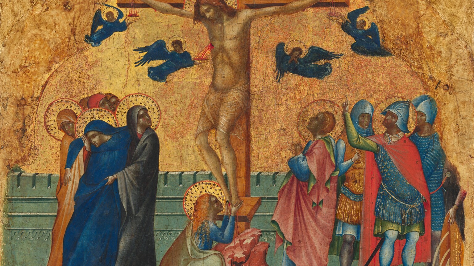 At least eleven people gather around a man hanging from a wooden cross, all against a gold background in this nearly square painting. The people have pale skin tinged heavily with gray, and pink on the cheeks. At the center of the composition, the man, Jesus, hangs from nails driven into the cross through his palms and feet. His thin body is covered only by a sheer loincloth across his hips. His knees angle to our left, and his head tips in that direction. He has a beard and auburn-brown, shoulder-length hair. His eyes are closed under furrowed brows. A gold halo surrounds his head and has red lines radiating to the red outline of the disk. The letters “INRI” appear on a panel on top of the cross. Three small, winged angels, like miniature people, catch vibrant red blood spurting or trickling from Jesus’s two palms and a gash over his right ribs in gold bowls. A fourth angel clasps hands in prayer. The angels have brown or blond hair, gold halos, and cobalt-blue robes and wings. A woman with long, curly, copper-colored hair kneels at the base of the cross, holding Jesus’s bleeding feet. To our left, a woman wearing lapis-blue swoons into the arms of two women who wear hoods and robes in marigold orange or nickel gray. All four women have gold halos, as do two of the four men standing together to our right. One of those men has short, copper-blond hair and wears a blue robe. His cloak is rose pink on one side and green on the other. He holds his small hands together in prayer and looks up at Jesus in profile. The other three men wear helmets and armored breastplates. One of them, at the front of that trio, has a halo and he points up at Jesus. The hand and torso of another person is visible along the right edge of this group, but the upper body has been lost. The cross sits on a low, rocky rise over a small cavern holding a skull. The ground around the cross and groups is carnation pink. A crenelated, turquoise-colored wall, about elbow height, runs across the back of the scene. The glimmering gold background is worn in some areas, revealing the red ground beneath the gold leaf.
