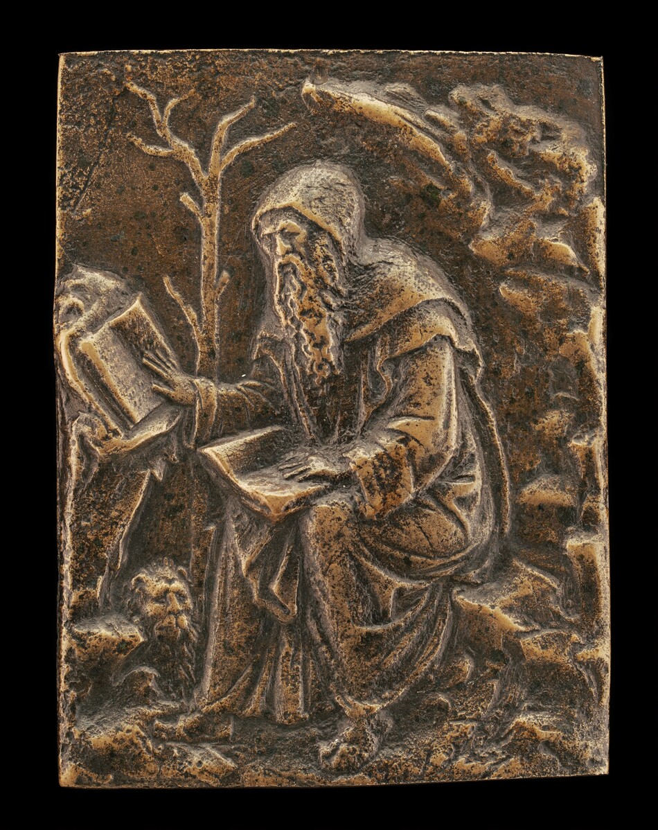 The sculpture is a relief depicting a seated figure wearing a hooded robe. The figure has a long beard and holds an open book in one hand, while looking at another open book on his knee. There is a bare tree behind him to the left and a rocky formation to the right. The sculpture is made of bronze, giving it a brownish-gold color with darker patina in the crevices. The composition is within a rectangular frame.