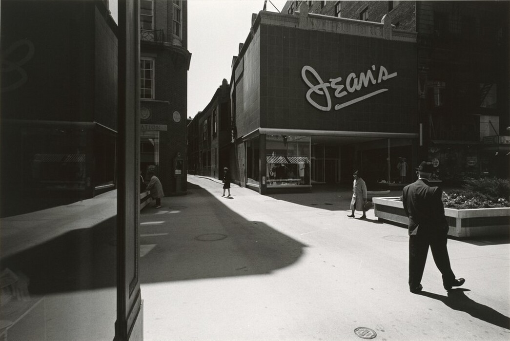 This is a photograph of a city street scene with a storefront featuring the word "Jean's" in large script letters. The storefront is part of a building in a shaded alleyway. The photograph emphasizes the contrasts between light and shadow, highlighting geometric shapes created by the buildings and pavement. Reflections from building windows add layers of light and dark. Display windows show mannequins or items.