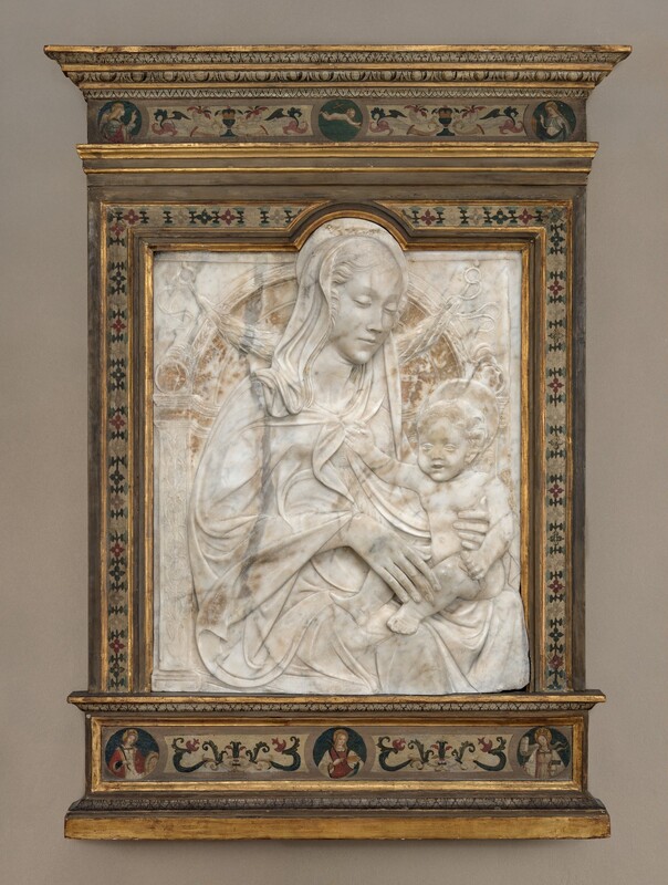 A woman and child are carved in low relief from a white marble, vertically oriented, rectangular panel, which is set within a painted wood frame. The woman’s body is angled to our right, and she looks down in that direction at the child seated on her lap. She has a long, delicate nose, a pointed chin, and her lips are closed. Her wavy hair is pulled back under a cloth that covers her heads and drapes over her shoulders. Her cloak waves in lively but shallow folds across the surface of the marble. She rests the long fingers of one hand on the baby’s shin and braces his side torso with the other. The nude boy has short, curly hair, round cheeks, chubby proportions, and his lips are parted. He holds a cylindrical object in one hand and reaches his right hand, to our left, to touch the woman’s clothing near her neckline. They both have flat, disk-like halos. The background is lightly carved to resemble an arched niche with leafy and geometric patterns and a leafy garland hanging from loops at the top corners. The marble panel is rounded at the top to encompass the woman’s halo and the top of her head. The frame is painted with stylized flowers and patterns in dark red, sky blue, ivory white, and black against a muted white background. Some of the stepped molding in the frame is gilded. A band across the top of the frame has roundels with two haloed women to either side and a nude baby carrying a cross over one shoulder, appearing to float against a blue background. Three roundels in a band below show women with long hair and halos, each holding objects like palm fronds, a plate, wheel, and a book.