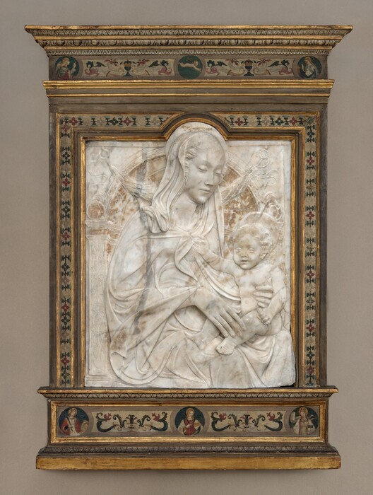 A woman and child are carved in low relief from a white marble, vertically oriented, rectangular panel, which is set within a painted wood frame. The woman’s body is angled to our right, and she looks down in that direction at the child seated on her lap. She has a long, delicate nose, a pointed chin, and her lips are closed. Her wavy hair is pulled back under a cloth that covers her heads and drapes over her shoulders. Her cloak waves in lively but shallow folds across the surface of the marble. She rests the long fingers of one hand on the baby’s shin and braces his side torso with the other. The nude boy has short, curly hair, round cheeks, chubby proportions, and his lips are parted. He holds a cylindrical object in one hand and reaches his right hand, to our left, to touch the woman’s clothing near her neckline. They both have flat, disk-like halos. The background is lightly carved to resemble an arched niche with leafy and geometric patterns and a leafy garland hanging from loops at the top corners. The marble panel is rounded at the top to encompass the woman’s halo and the top of her head. The frame is painted with stylized flowers and patterns in dark red, sky blue, ivory white, and black against a muted white background. Some of the stepped molding in the frame is gilded. A band across the top of the frame has roundels with two haloed women to either side and a nude baby carrying a cross over one shoulder, appearing to float against a blue background. Three roundels in a band below show women with long hair and halos, each holding objects like palm fronds, a plate, wheel, and a book.