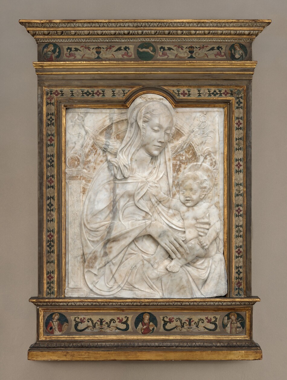 A woman and child are carved in low relief from a white marble, vertically oriented, rectangular panel, which is set within a painted wood frame. The woman’s body is angled to our right, and she looks down in that direction at the child seated on her lap. She has a long, delicate nose, a pointed chin, and her lips are closed. Her wavy hair is pulled back under a cloth that covers her heads and drapes over her shoulders. Her cloak waves in lively but shallow folds across the surface of the marble. She rests the long fingers of one hand on the baby’s shin and braces his side torso with the other. The nude boy has short, curly hair, round cheeks, chubby proportions, and his lips are parted. He holds a cylindrical object in one hand and reaches his right hand, to our left, to touch the woman’s clothing near her neckline. They both have flat, disk-like halos. The background is lightly carved to resemble an arched niche with leafy and geometric patterns and a leafy garland hanging from loops at the top corners. The marble panel is rounded at the top to encompass the woman’s halo and the top of her head. The frame is painted with stylized flowers and patterns in dark red, sky blue, ivory white, and black against a muted white background. Some of the stepped molding in the frame is gilded. A band across the top of the frame has roundels with two haloed women to either side and a nude baby carrying a cross over one shoulder, appearing to float against a blue background. Three roundels in a band below show women with long hair and halos, each holding objects like palm fronds, a plate, wheel, and a book.