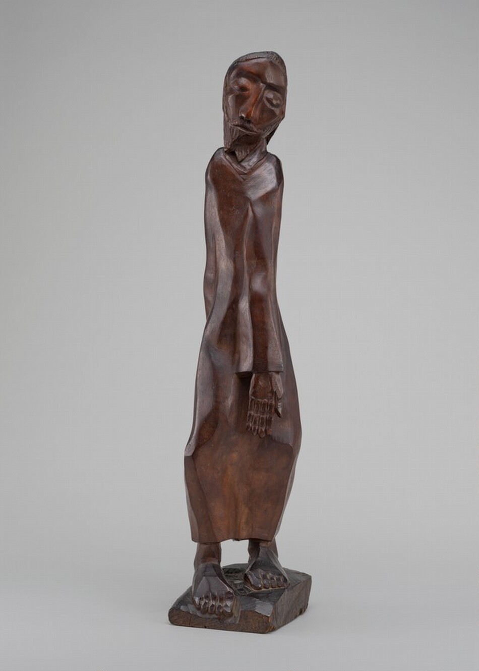 The sculpture depicts a human figure with a slightly tilted head and closed eyes. The figure is carved from wood, showcasing a rich, dark brown color with a smooth, polished surface that highlights the natural grain of the material. The person wears a long, flowing garment that drapes down to the base, with feet positioned flat on a rectangular platform. The sculpture is abstract, with stylized facial features and limbs, giving it a somewhat elongated and angular appearance.