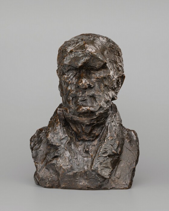 The image depicts a bust sculpture focusing on the head and shoulders of a human figure. The sculpture is made of bronze, showing rough textures on both the facial features and clothing. The plain background enhances the prominent features of the sculpture.