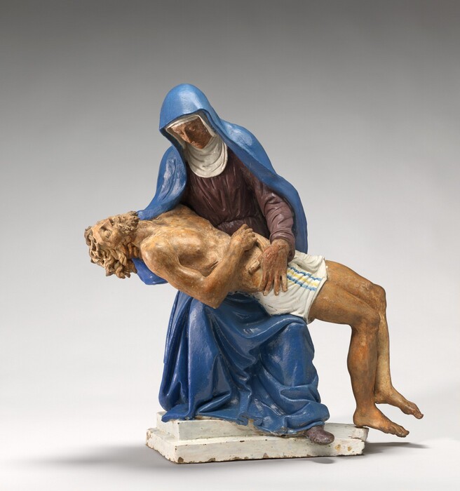 A woman braces the body of a dead young man across her lap in this freestanding, painted, terracotta sculpture. The woman half stands and half crouches with her body facing us in this photograph. Her face is mottled with red and peach and is lined, and her eyes look down or are closed. Her snow-white head covering also lines her face, covers her neck, and rests in folds on her chest. Her lapis-blue cloak drapes over her head and falls to her feet over her long-sleeved, plum-purple dress. The man’s skin is glazed with mottled shades of tan and peach, and he wears only a white loincloth across his hips. The woman supports his head with her right hand, to our left, so her fingers twine with his long curls. The man has a curly beard. His lips are parted, and his eyes are almost closed. His right arm, closer to us, lies across his belly and there is a hole piercing the back of that hand. There is also a gash across his right ribs and holes in the tops of his feet. The woman braces his hip with her other hand, and his legs fall limply to the side, knees bent. The woman stands on a step on a white base, and one purple shoe peeks out under the pooling folds of her blue cloak. Some of the painted, glazed areas are chipped on the white base.